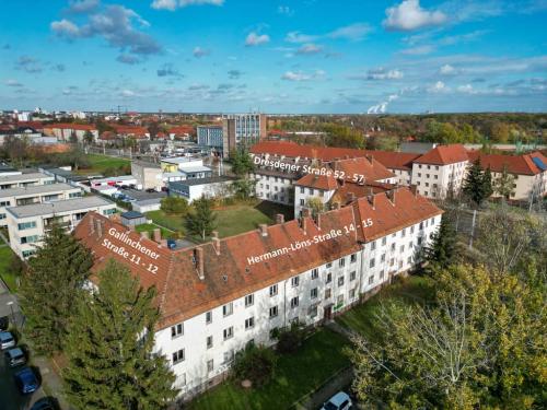 Das Bild zeigt eine Luftaufnahme eines Wohngebiets. Im Vordergrund stehen mehrere mehrstöckige Wohnhäuser mit roten Ziegeldächern und hellen Fassaden. Im Hintergrund sind weitere Gebäude, Grünflächen und ein Teil der Stadtlandschaft zu sehen. Der Himmel ist klar und hellblau. Das Bild wirkt insgesamt ruhig und zeigt eine geordnete Bebauung.