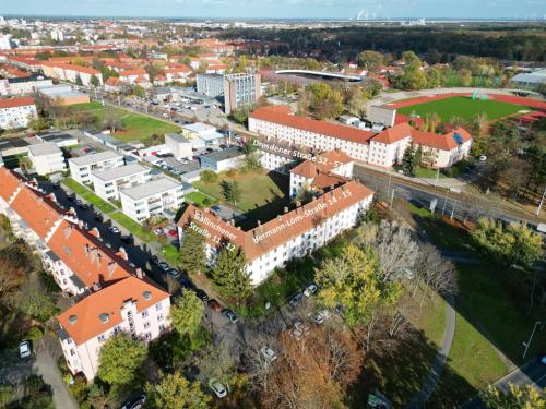 Das Bild zeigt eine Luftaufnahme eines Wohngebiets. Im Vordergrund stehen mehrere mehrstöckige Wohnhäuser mit roten Ziegeldächern und hellen Fassaden. Im Hintergrund sind weitere Gebäude, Grünflächen und ein Teil der Stadtlandschaft zu sehen. Der Himmel ist klar und hellblau. Das Bild wirkt insgesamt ruhig und zeigt eine geordnete Bebauung.